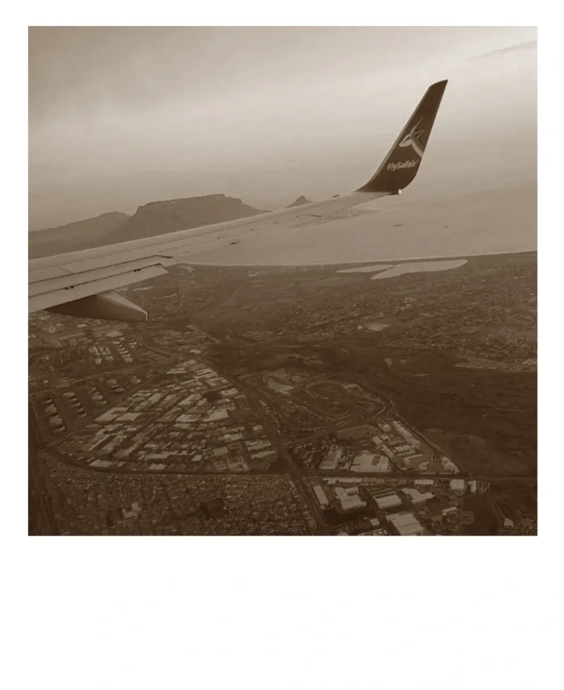 Fat Barrel Wine Company: A sepia-toned photograph taken from an airplane window, showing the wing of the plane with a view of the landscape below. The scene captures the excitement and anticipation of arriving at a new destination, with the horizon stretching out in the distance.